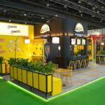 Bright exhibition booth with yellow walls labeled 'Country Crinkers' and a McCain counter area with stools and plants nearby.