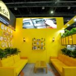 Bright yellow exhibition booth featuring yellow couches, green plants, and a central coffee table under warm lighting, with a McCain sign above left.