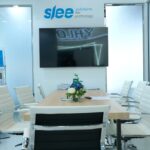 Modern conference room with a wooden table, white leather chairs, and a wall-mounted TV; flowers centerpiece and glass walls.