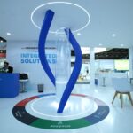 Blue twisted sculpture with a circular glowing ring atop a white showroom floor at a trade show booth labeled 'Integrated Solutions'.