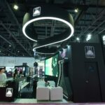 World vape show exhibition stand by Spectrum Exhibition - Spectrum Monster Vape Labs exhibit booth with a circular glowing ring overhead, black curved walls, a large video screen, and white lounge seating at a convention hall.