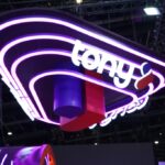 Purple neon sign hanging from a ceiling, displaying the white word 'toy' in a rounded rectangular frame at an exhibition space.