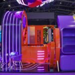 Trade show booth with orange reception counter, purple shelving, and neon-lit curved arches.